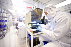 A person in a white cleanroom suit and blue gloves operates machinery in a bright laboratory.