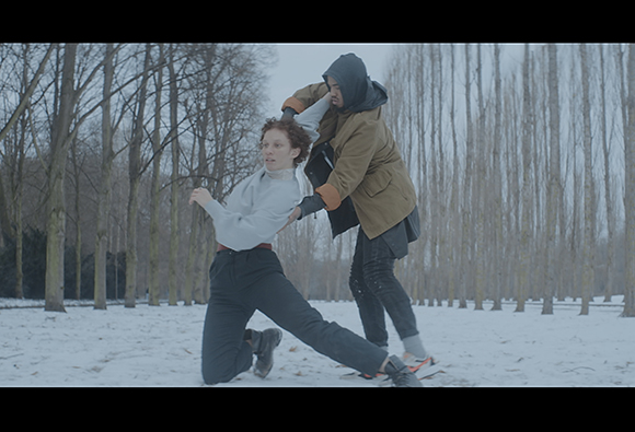 A person in a light sweater is being held by another person in a brown jacket in a snowy wooded area.