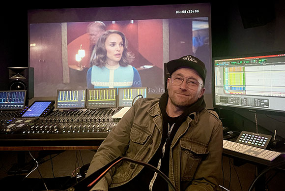 A man in a cap and glasses sits in a dark studio before a large mixing console and video screens.