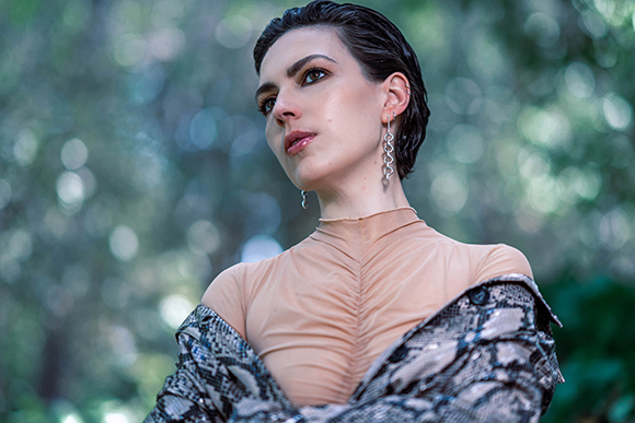 A woman with dark hair wearing a tan turtleneck and a snakeskin patterned wrap in a forest.
