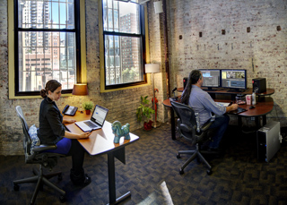A woman uses a laptop at a desk while a man works with dual monitors at a curved desk in a brick office.