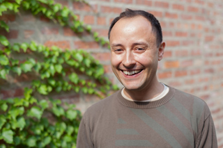 A smiling man wearing a brown sweater stands in front of a brick wall covered in green ivy.