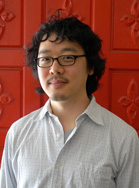 A man with dark curly hair and glasses wearing a light grey button-down shirt against a red patterned door.