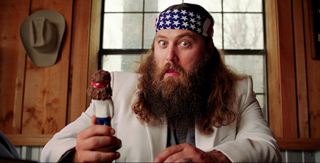 A man with a long beard and star-patterned headband holds a small figurine in a wooden room.