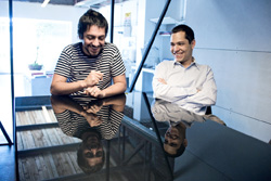Two smiling men sit at a dark, reflective table in a bright office.