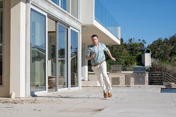 A man in a patterned shirt and white pants jumps in the air on a stone patio.