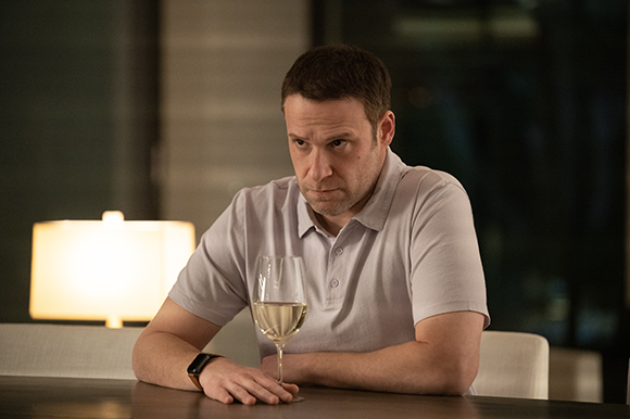 A man in a light polo shirt sits at a table looking contemplative next to a glass of white wine.