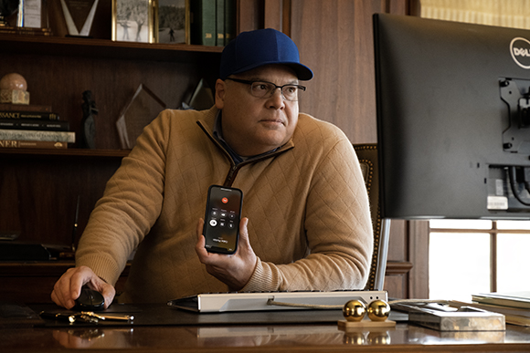 A man in a blue cap and tan sweater looks at a computer screen while holding a smartphone in his hand.