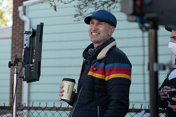 A smiling man in a blue cap and black puffer jacket holds a coffee cup outdoors.