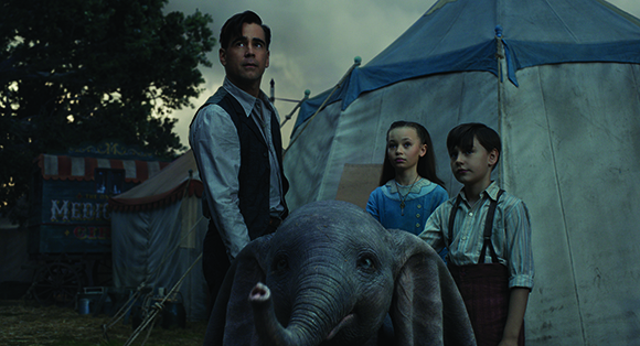 A man in a vest and two children stand beside a small elephant in front of a large circus tent.