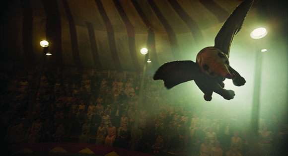 A large, winged creature flies through a misty circus tent above a seated audience.