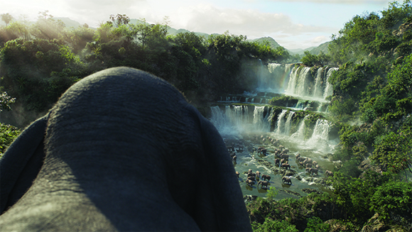 View from behind an elephant's head looking over a tropical valley with large waterfalls and a herd of elephants.
