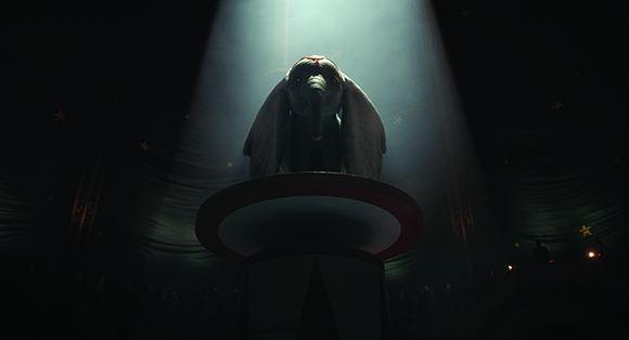 An elephant stands on a circular pedestal under a single bright spotlight inside a dark circus tent.