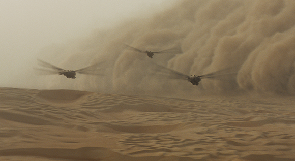 Two helicopters fly through a large, billowing sandstorm over desert sand dunes.