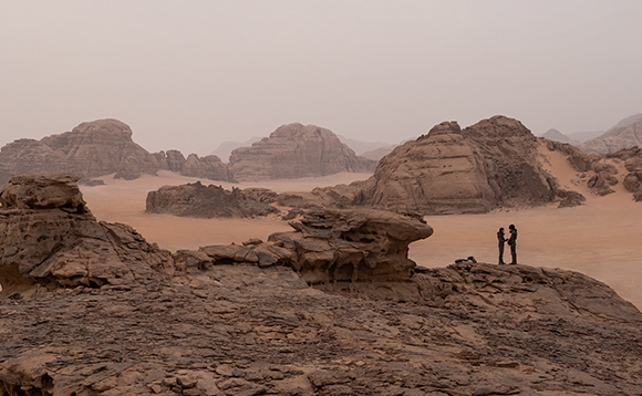 Two people stand facing each other on a large rock formation in a vast desert with mountains.
