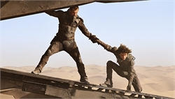 A man in dark tactical gear pulls another person up a metal ramp in a vast desert.