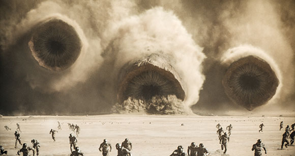 Three giant sandworms emerge from clouds of dust in a desert as people run across the sand below.
