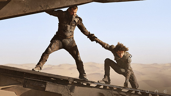 A man in dark tactical gear reaches down to pull a woman up a metal ramp in a vast desert.