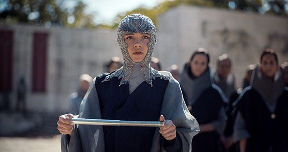 A young person wearing a silver textured headpiece and dark robes holds a silver rod in front of a group.