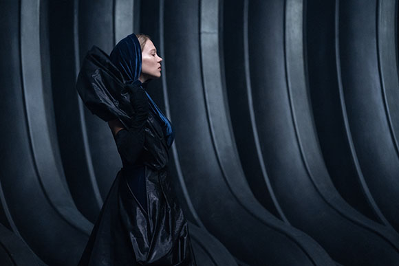 Profile of a person wearing a voluminous black garment and blue headscarf against dark, curved architectural lines.