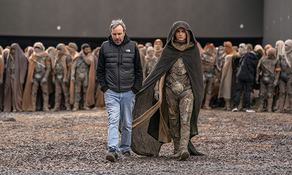 A man in a black puffer jacket walks next to an actor in a hooded desert cloak on a gravel set.