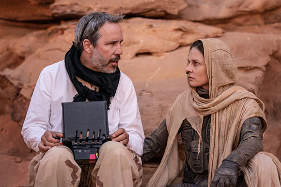 A man in a white shirt and black scarf talks to a woman wearing a tan headscarf in a desert setting.