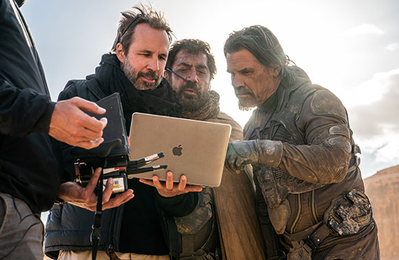 A man in a black jacket holds a laptop while a man in costume and another crew member look at the screen.