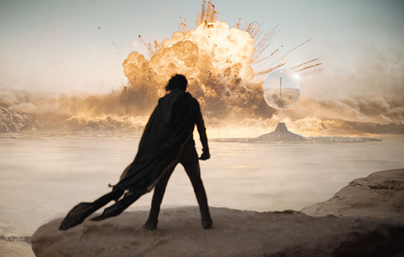 Silhouette of a person in a cape watching a large explosion and a glass sphere over water.