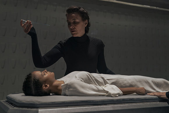 A woman in a black turtleneck stands over a person lying on a gray table in a dimly lit room.