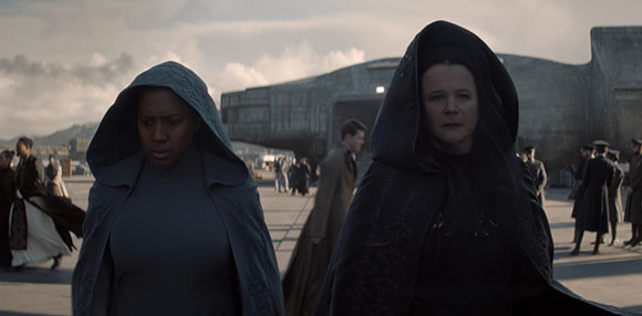 Two women wearing dark, heavy hooded cloaks walk through an outdoor area near a large concrete structure.