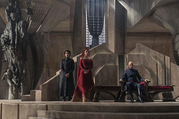 Three people in dark and red formal clothing pose in a massive, geometric stone hall with a large statue.