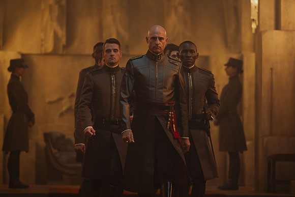 Four men in dark, structured military uniforms stand in a dimly lit, stone hall.