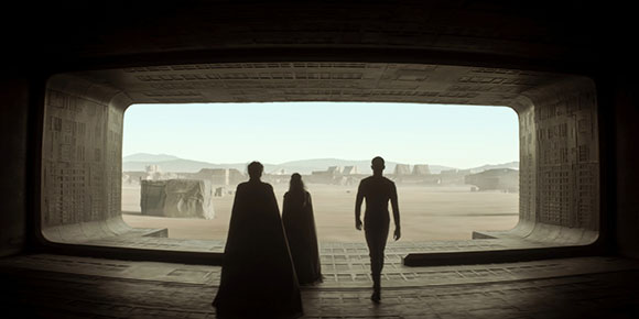 Silhouettes of three people standing in a dark doorway looking out at a vast, sandy desert landscape.