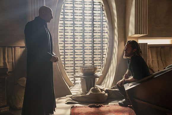 A man in a long black coat stands before a seated man in a room with a large, patterned circular window.