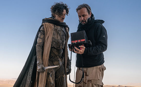 A man in detailed sci-fi armor and a cape stands beside a man reviewing footage on a handheld monitor.