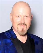 A bald man with a light brown goatee wearing a blue patterned blazer over a black shirt.