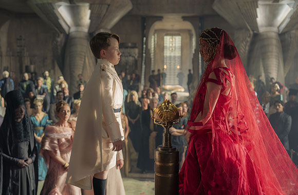 A young boy in a white cape faces a woman in a red gown and veil in a large, dimly lit hall.