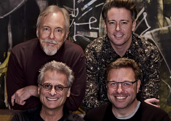 Four smiling men posing closely together against a dark, abstract background.