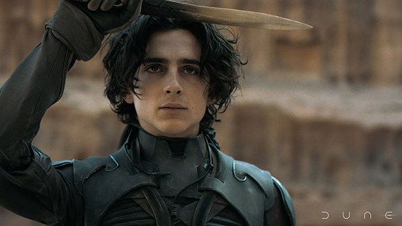 A man with dark curly hair wearing dark tactical armor holds a long blade above his head.