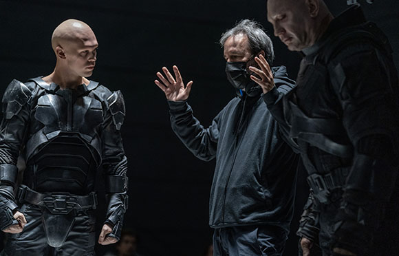 A man in a black hoodie gestures with raised hands toward two men wearing black armored suits.