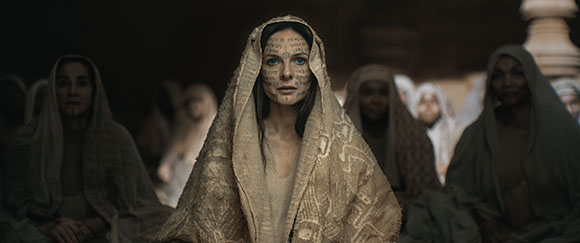A woman with blue eyes and intricate facial markings wears a light-colored patterned head covering.