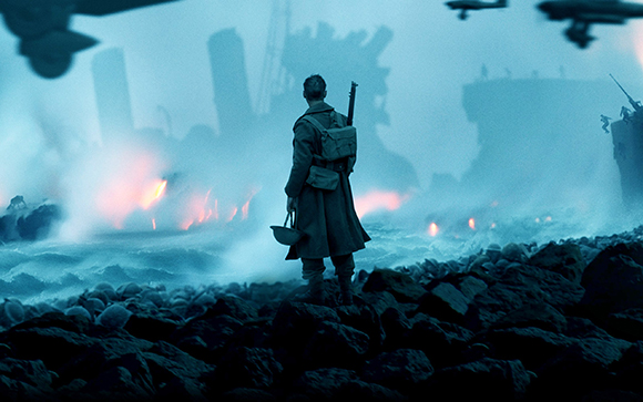 A soldier in a long coat stands on dark rocks facing a blue, misty shoreline with distant silhouettes and fires.