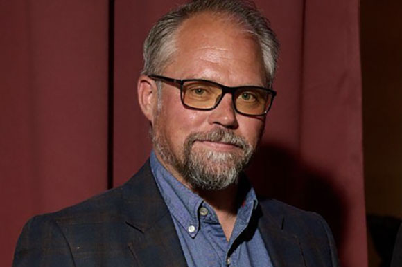 A man with short grey hair, glasses, and a salt-and-pepper beard wearing a blue shirt and dark blazer.