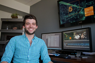 A smiling man in a blue checkered shirt sits at a desk with two computer monitors and a large screen.