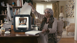 Elderly woman with glasses and a leopard print scarf lifting the lid of a small white device on a desk.
