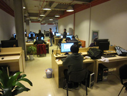 Several people sit at desks with computer monitors in a brightly lit office or computer lab.