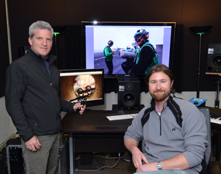 Two men pose in a studio with large monitors displaying video footage and studio speakers.
