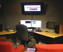 A video editing desk with two computer monitors, a large wall-mounted TV, and various electronic devices.