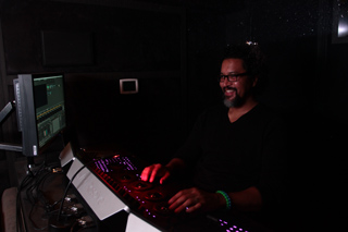 A man with glasses and a beard smiles while operating a lit audio mixing console in a dark room.