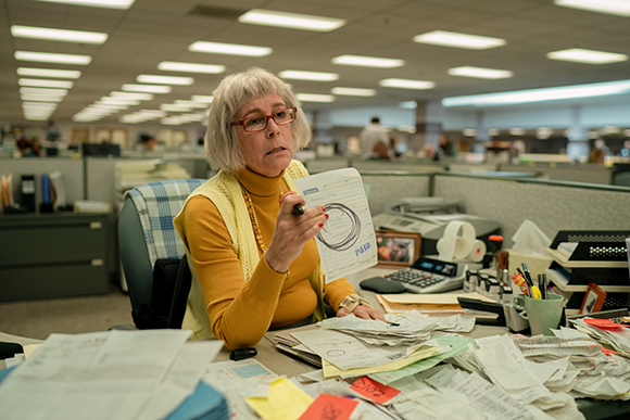 An older woman with glasses holds up a paper document in a busy, cubicle-filled office.